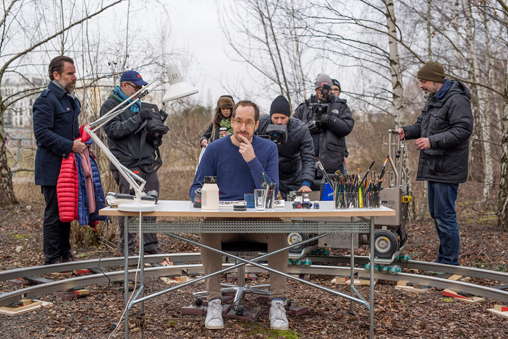 Ein Mann sitzt an einem Zecihentisch in der Natur, um ihm herum ein Kamerateam