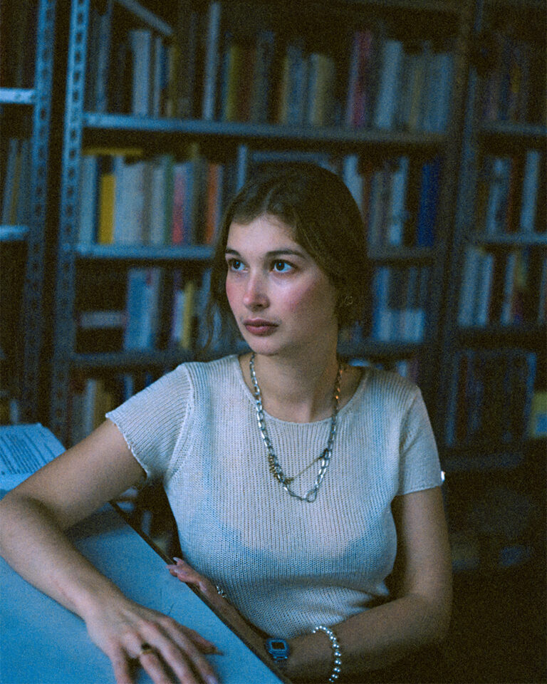 Die Autorin Olga Hohman sitzt vor einer Bücherwand an einem Schreibtisch und hat ihren Arm darauf abgelegt. Sie hat ihren Kopf zur Seite gedreht und blickt in diese Richtung.